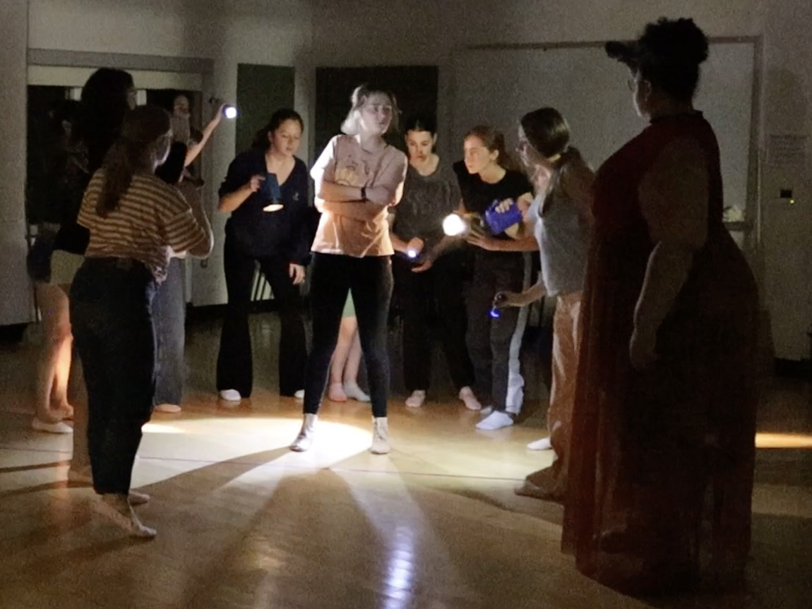 Photo of a group of college-age actors stands in a group, rehearsing. The lights in the room are low, and the group is lit by hand-held flashlights.